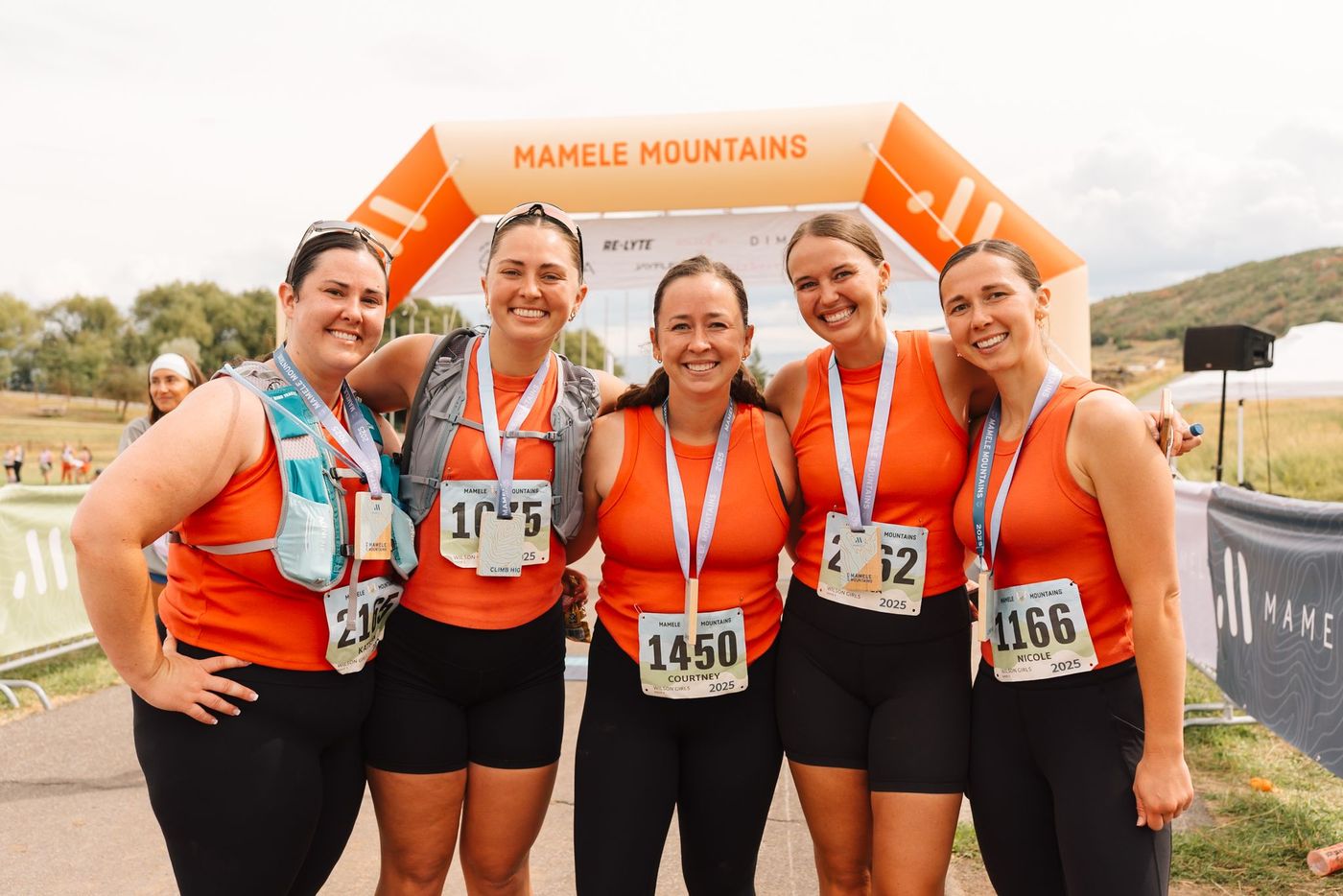 Five women with finisher medals standing under the Mamele Mountains arch.